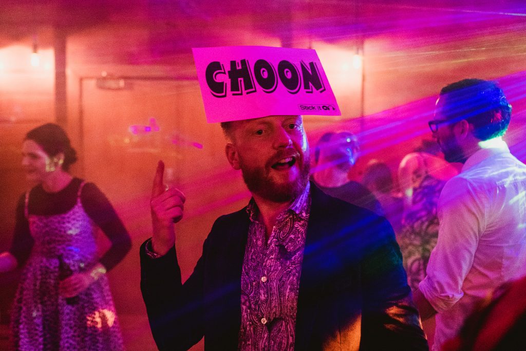 Guest dancing with ‘CHOON’ sign on head beneath colourful lights at wedding reception.