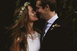 Bride and groom smiling nose to nose beneath greenery, intimate documentary wedding photography moment.