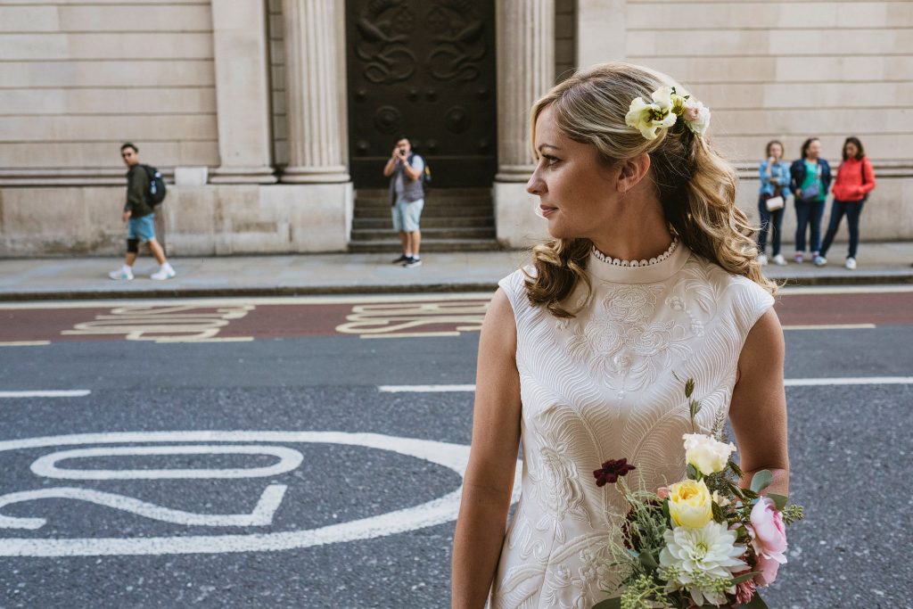 Bride holding bouquet on city street, glancing sideways as pedestrians pass, documentary wedding photography.