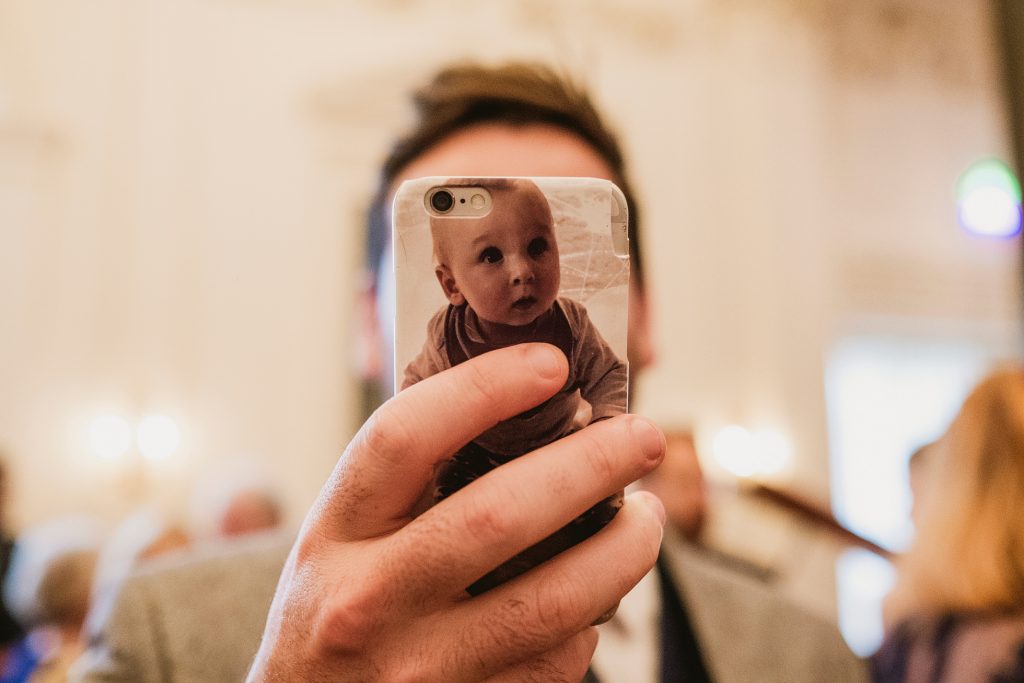Guest holds phone with baby photo case covering his face during wedding reception.