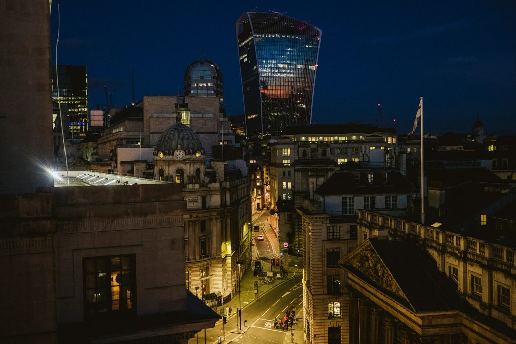 Twilight over London skyline, office windows glowing above quiet streets where a small group crosses under lamplight.