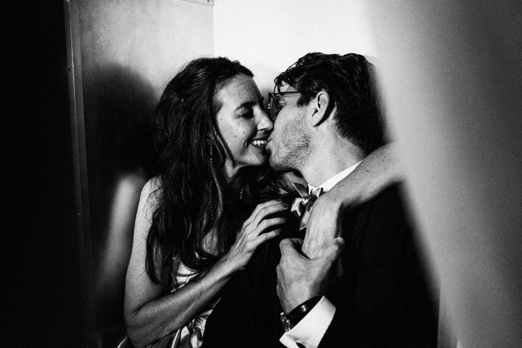 Bride and groom share a quiet kiss in shadowed corner, candid wedding photography moment.