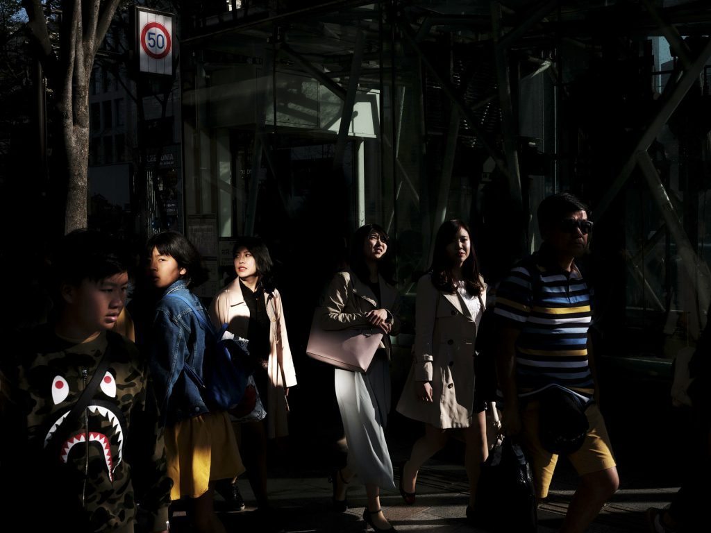 Commuters cross sunlit city street, faces briefly illuminated against deep evening shadows.