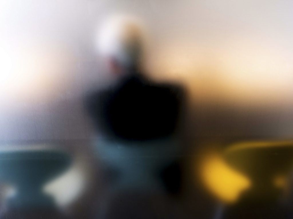 Softly blurred figure seated between teal and mustard cushions in warm indoor light.