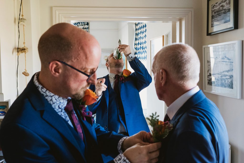 Groom drinks from a can as two men adjust buttonholes in a bright room on the wedding morning.