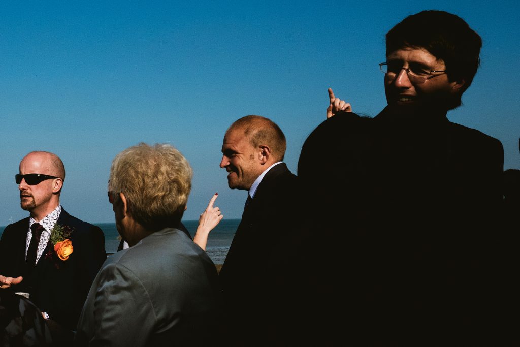 Wedding guests laugh and point by the sea under clear blue sky, candid documentary wedding photography moment.