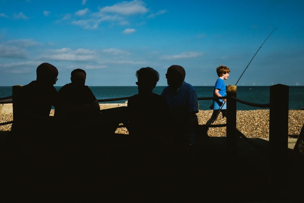 Silhouetted figures sit by the sea as a boy walks past holding a fishing rod.