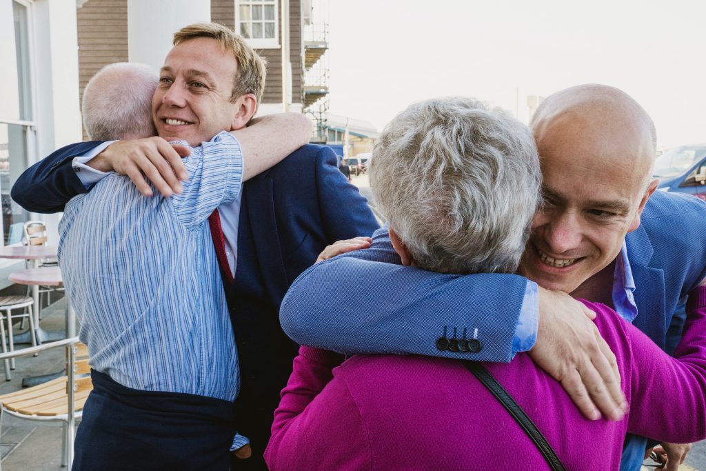 Two men in suits warmly embrace older relatives outside, smiling in bright afternoon light, wedding photography.