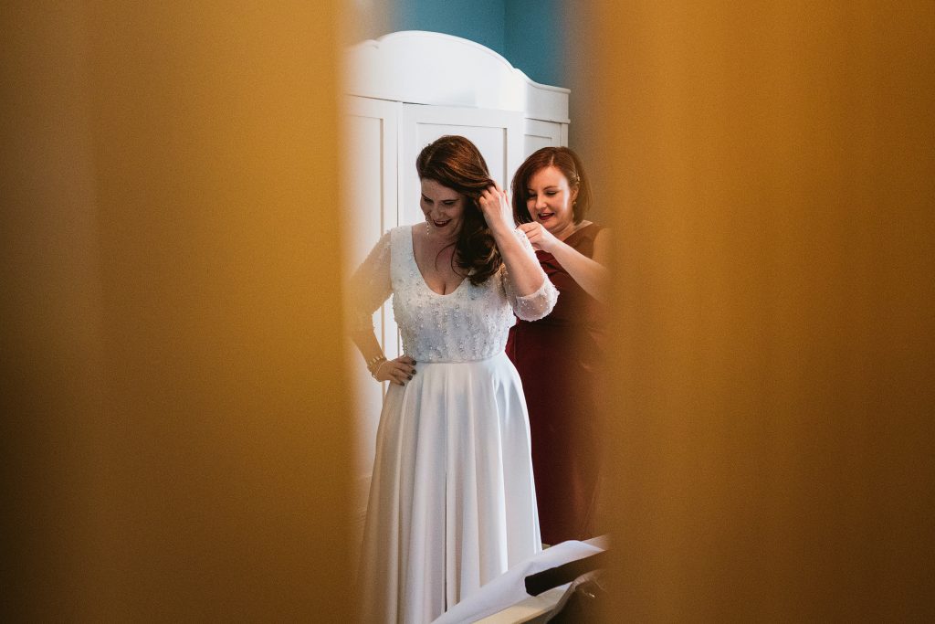 Bride smiles as friend fastens her dress in soft window light, documentary wedding photography moment.
