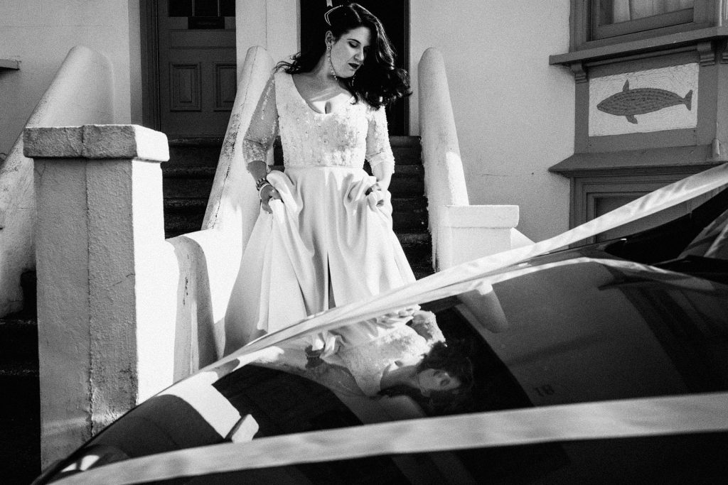Bride lifts her dress while stepping past a car, her reflection shimmering in the roof, in documentary wedding photography.