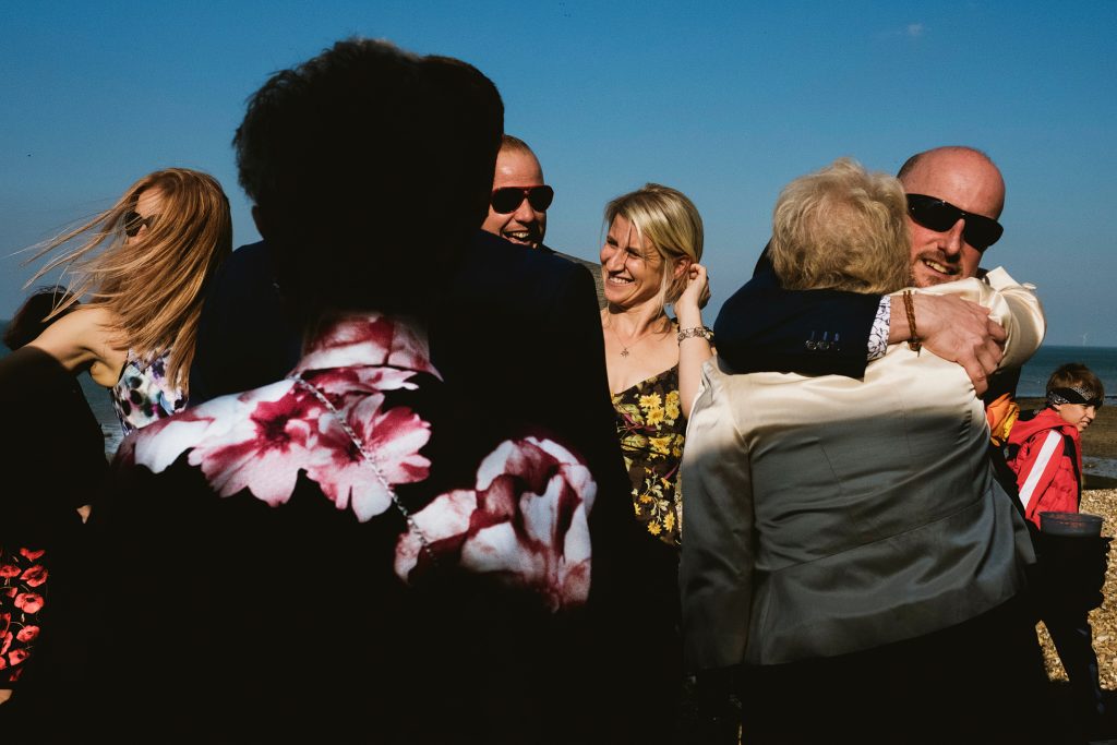 Guests laughing and embracing by the seaside during a sunlit wedding celebration.