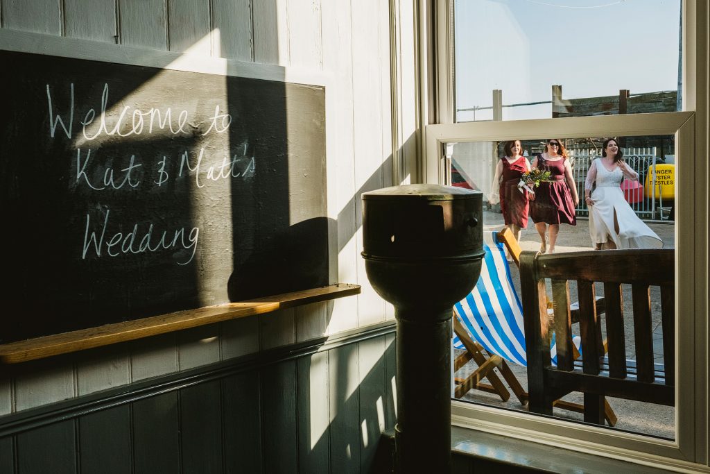 A documentary wedding photograph at the entrance of East Quay, Whitstable. The bride is captured walking into the venue, naturally framed by her bridesmaids and the iconic "East Quay" signage. The shot focuses on the unscripted anticipation of the arrival, using the venue’s external architecture to frame a raw, cinematic moment.