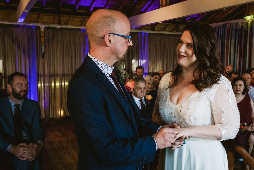 Couple hold hands and exchange smiles during intimate indoor wedding ceremony, guests watching softly, captured in documentary wedding photography.