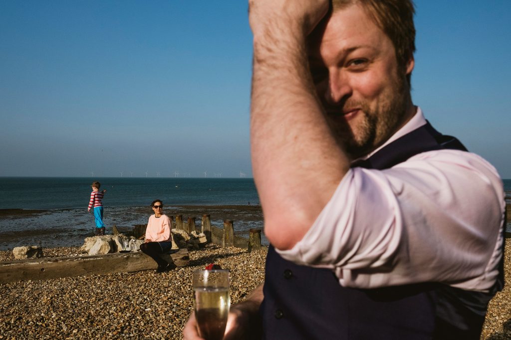 Man with champagne on a pebbled beach, woman and child by the sea in soft evening light.