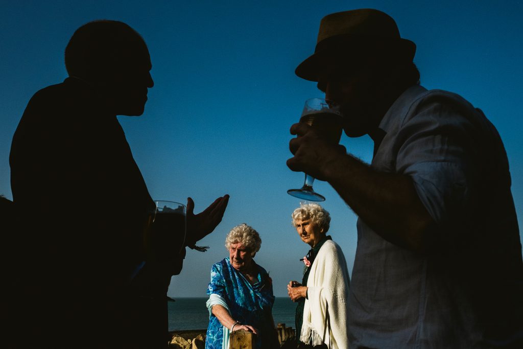 Silhouetted guests chat with drinks as two elderly women stand by the sea at a wedding.