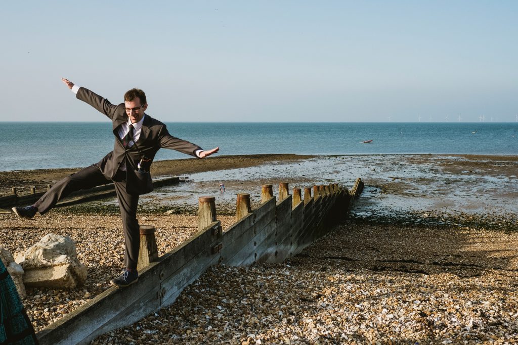 A candid documentary photograph on the Whitstable shingle beach at East Quay. The frame captures an adult guest messing around and playing on the pebbles, highlighting the unscripted and humorous side of the wedding day. The shot focuses on the "unfiltered" energy of the guests against the vast, open backdrop of the North Sea horizon.