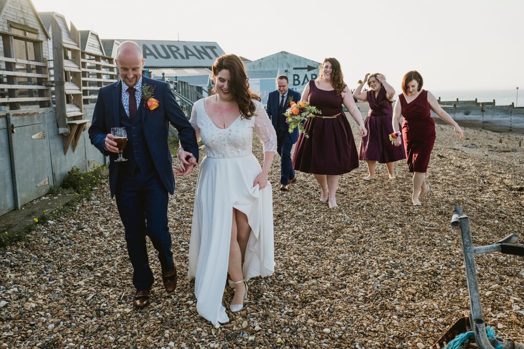 Unposed wedding photography at East Quay Venue, Whitstable. A cinematic study of light and shadow as the bridal party walks the pebble beach at sunset, emphasizing the "weathered charm" of the Kent coast and the honest human connections of the day.