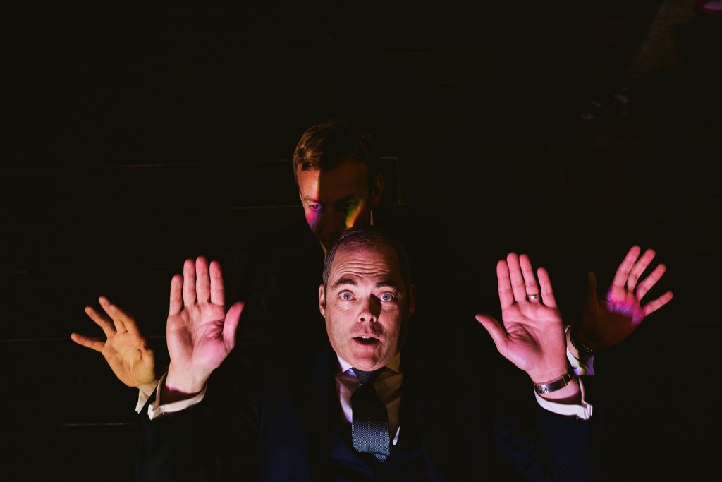 Two suited guests raise their hands under coloured lights at a wedding reception.