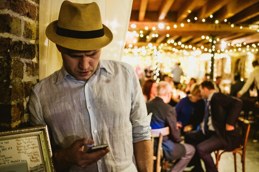 Guest in trilby checks his phone beside brick wall as fairy lights glow over wedding reception.