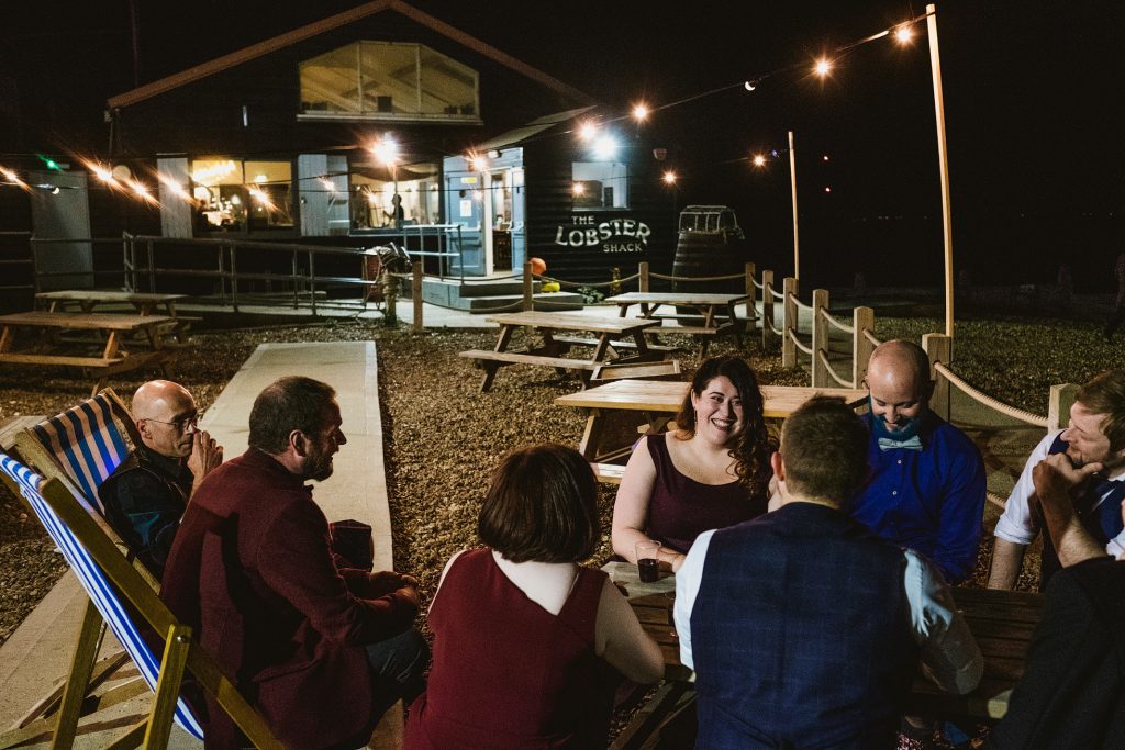 Guests laugh around picnic tables outside The Lobster Shack at night, fairy lights glowing overhead — wedding photography.