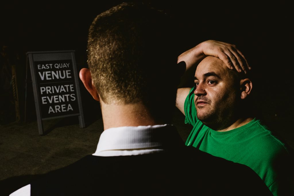 Two men talk in dim light beside East Quay wedding venue sign.