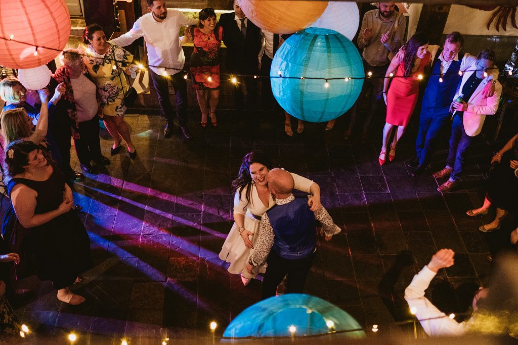 A cinematic high-angle shot from the East Quay mezzanine, capturing the first dance below. The composition highlights the industrial geometry of the room and the "Harbour Pulse" of the evening reception, looking down on the couple and their guests in a raw, unscripted moment of celebration.