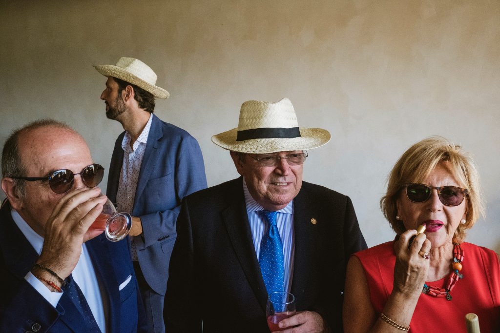 Wedding guests in summer hats sip drinks and apply lipstick against a plain wall.