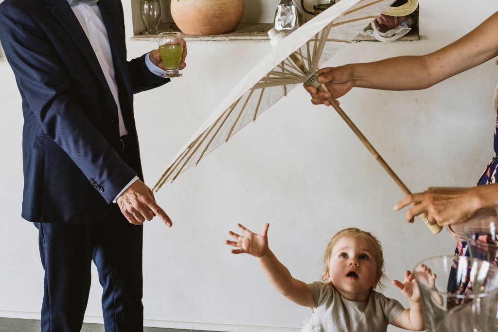 Toddler reaches towards open parasol as suited guest points, drink in hand, during wedding celebration.