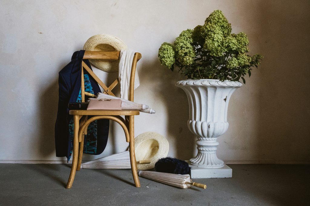 Jacket and straw hat draped over chair beside umbrellas and hydrangeas in stone urn, wedding morning stillness.