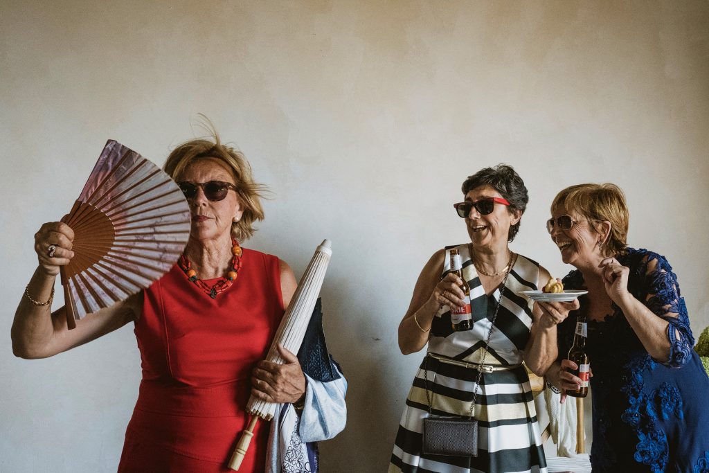 Three women in sunglasses laugh and cool themselves with fans and drinks at a summer wedding reception.