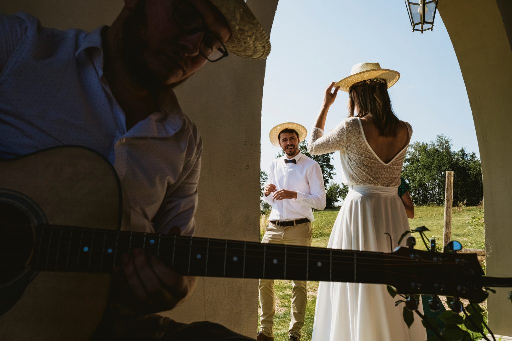 Bride adjusts straw hat as groom smiles in doorway, guitarist playing softly nearby during documentary wedding photography.