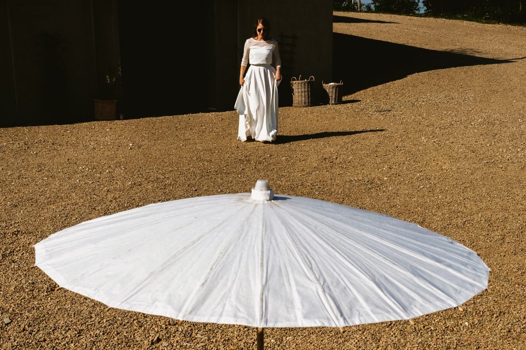 Bride walking across sunlit gravel, white parasol in foreground, quiet wedding morning light.
