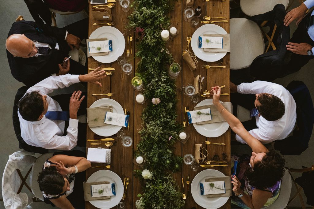 Overhead view of guests gathered at a wedding reception table with greenery centrepiece and golden cutlery.