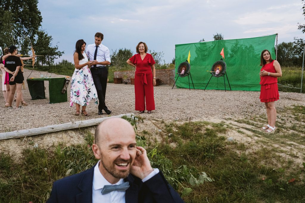 Guests gather beside outdoor archery targets as a smiling man steps forward, summer wedding evening.