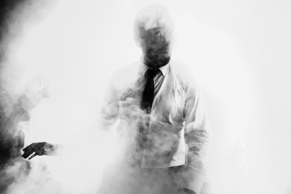 Groom in white shirt and black tie stands in drifting smoke, wedding photography in quiet monochrome.