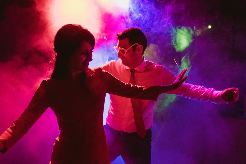 Couple dancing on wedding dancefloor beneath pink and green lights, laughing in drifting smoke.