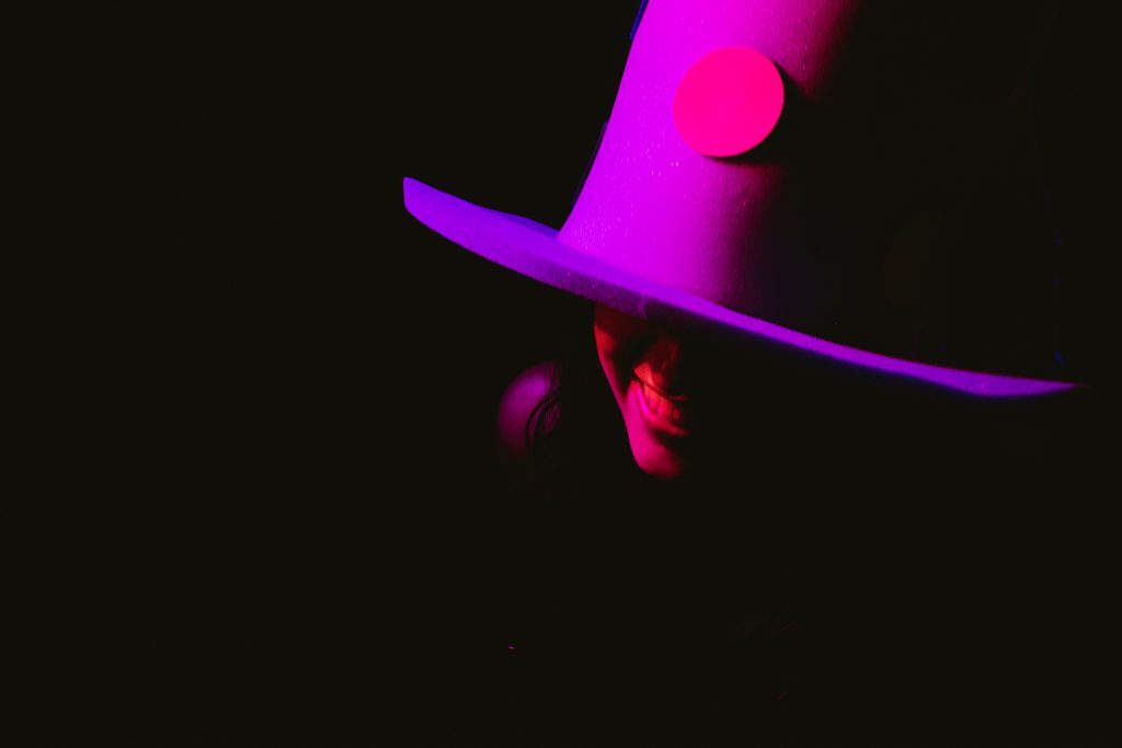 Guest in tall top hat smiles under pink light on dark dancefloor, wedding photography moment.