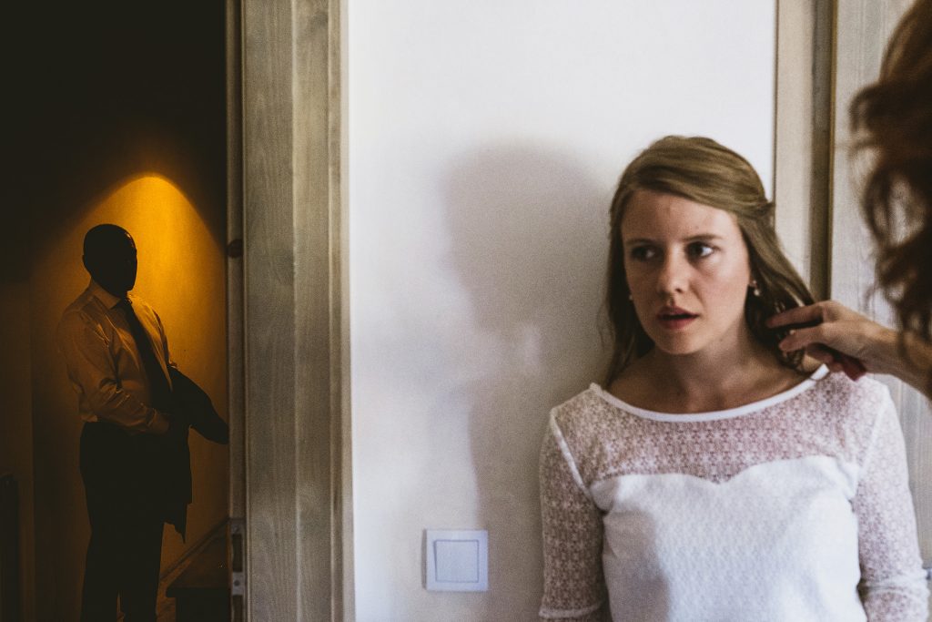 Bride leans against wall as hair is adjusted, a figure waits in warmly lit doorway — documentary wedding photography