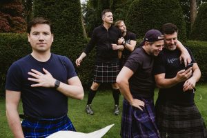 Friends in kilts laugh together in a leafy garden, one reading a phone, during relaxed wedding photography.