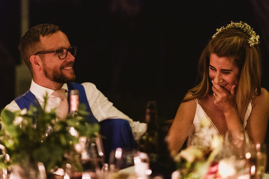Bride laughs with hand over mouth as groom smiles across candlelit table during wedding speeches