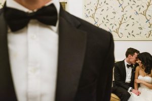 Bride and groom laughing together on sofa, partially framed by tuxedo jacket, intimate wedding photography moment.