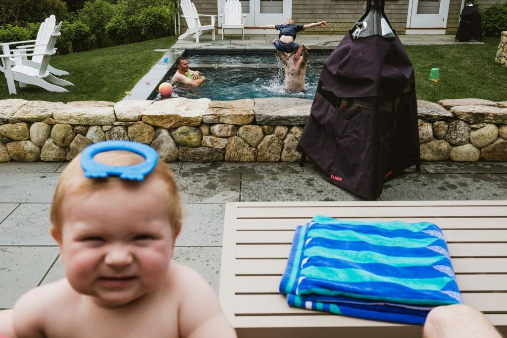 Baby with blue toy on head watches adults splash in garden pool during wedding weekend.