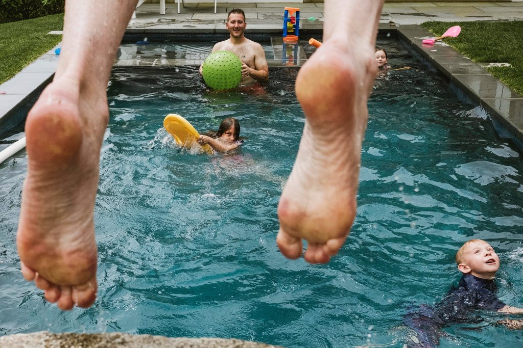 Bare feet leap into a garden pool as children splash and a man holds a green ball.