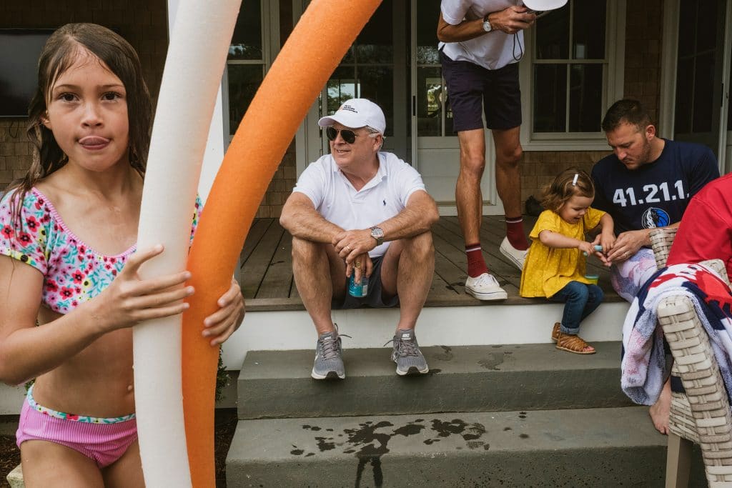 Girl in swimsuit holds pool noodles as family sit chatting on sunlit porch steps.
