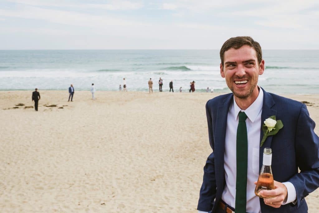 Groom laughing on sandy beach, holding drink as waves roll behind during beach wedding.