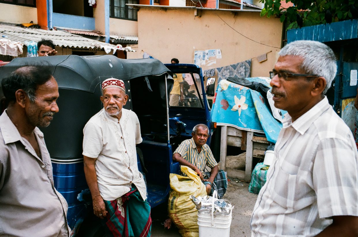 Four men chatting beside a blue tuk-tuk on a quiet street, everyday life unfolding in warm afternoon light.