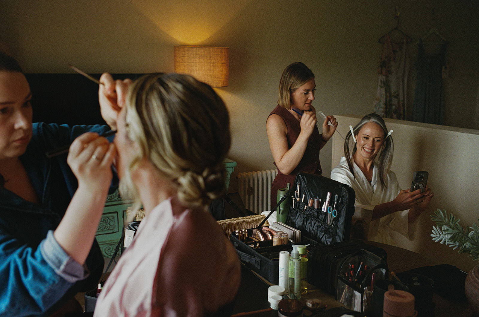 Bride smiling at her phone as stylists curl her hair on a softly lit wedding morning.