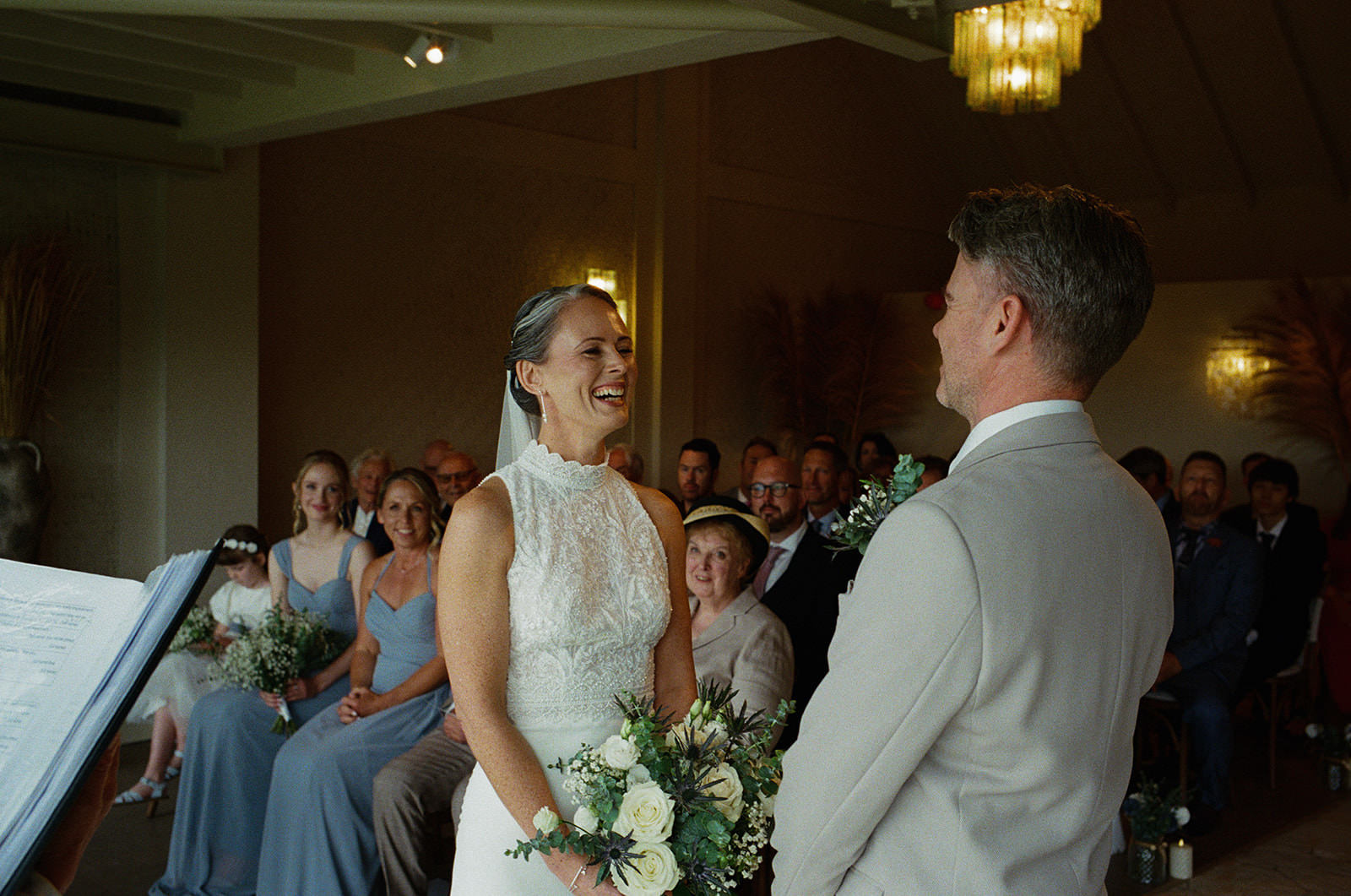 Bride laughing during intimate wedding ceremony as guests watch, holding white rose bouquet.