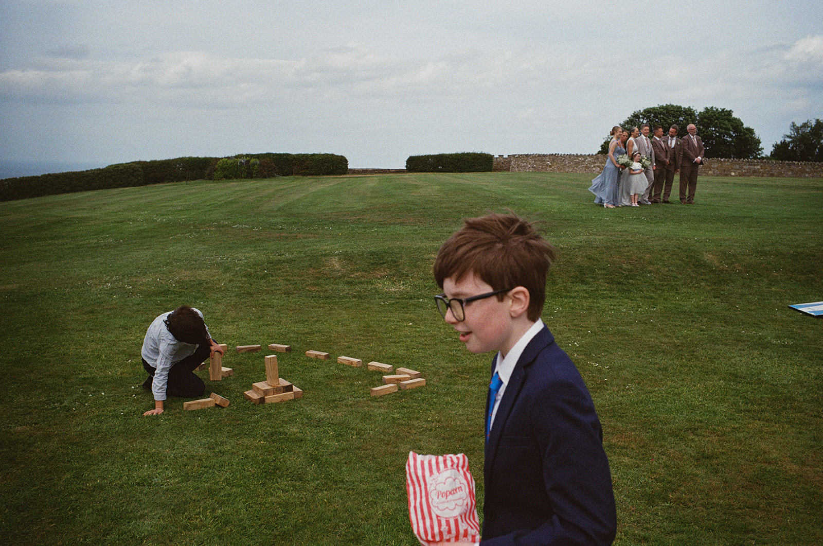 Boy in suit walks across lawn with popcorn as children play giant Jenga at outdoor wedding.