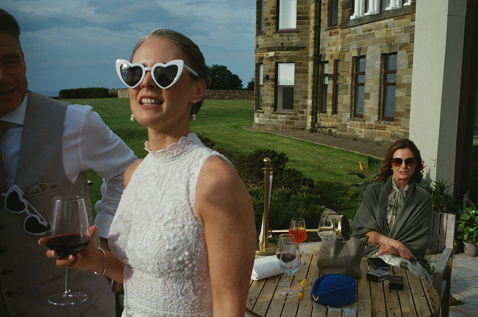 Bride in heart-shaped sunglasses holds wine, laughing on lawn outside stone house during wedding reception.